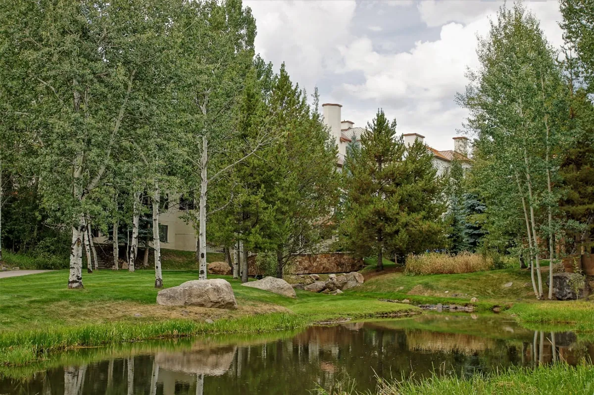 A reflective pond surrounded by tall trees, rock formations, and a building in the backdrop. The scene is serene, inviting guests to stroll, walk, or simply take in the natural beauty, representative of the peaceful neighborhood setting.