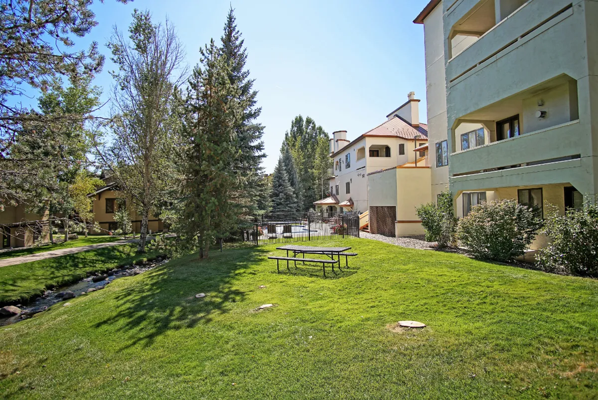 This exterior image depicts a cheerful picnic area with a picnic table set on a lawn, surrounded by trees and with the building's facade in the background. The setting is bright and inviting, ideal for outdoor gatherings and creekside BBQs.