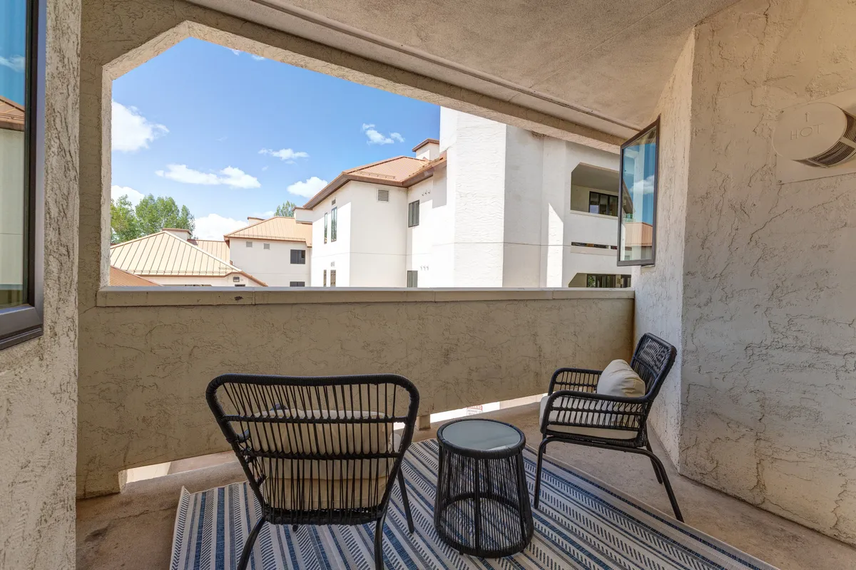 This image features two black chairs and a small round table set on a tranquil balcony with a striped rug and a view of nearby buildings. The setting evokes intimacy and relaxation, perfect for unwinding with a drink while enjoying sunsets.