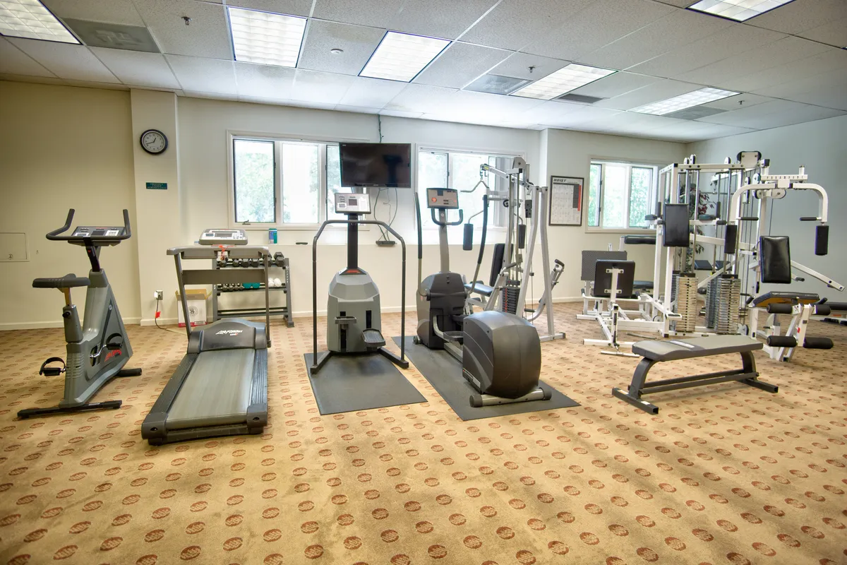 This interior image features a well-equipped fitness center with cardio machines such as a treadmill, elliptical machine, stationary bike, and a weight machine. A large television is mounted for entertainment, and the space appears clean, modern, and motivating.