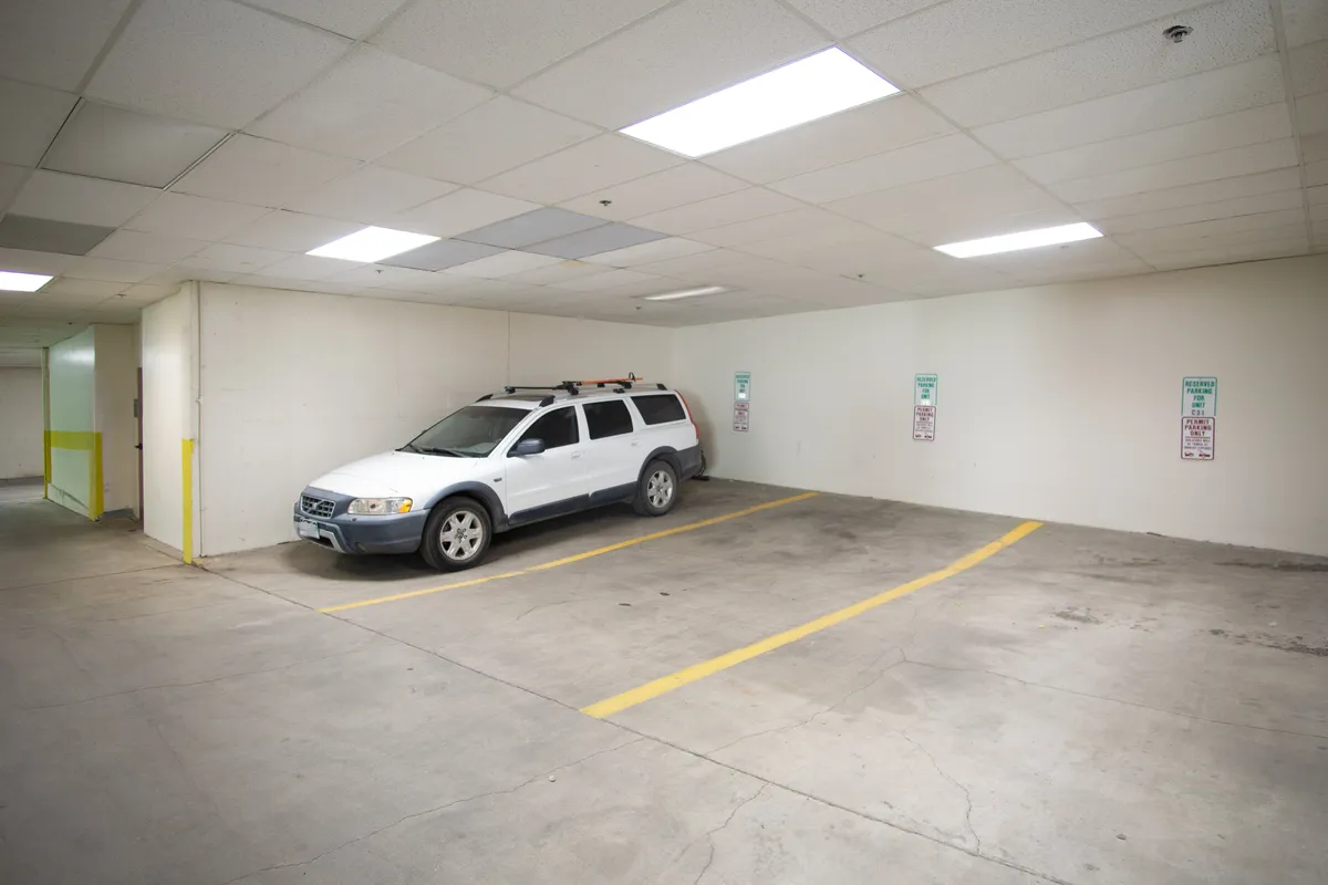 A white SUV is parked in a heated underground parking garage, with empty spaces and clear wall signage visible. The area looks secure and spacious, emphasizing practicality and comfort for long-term guests.