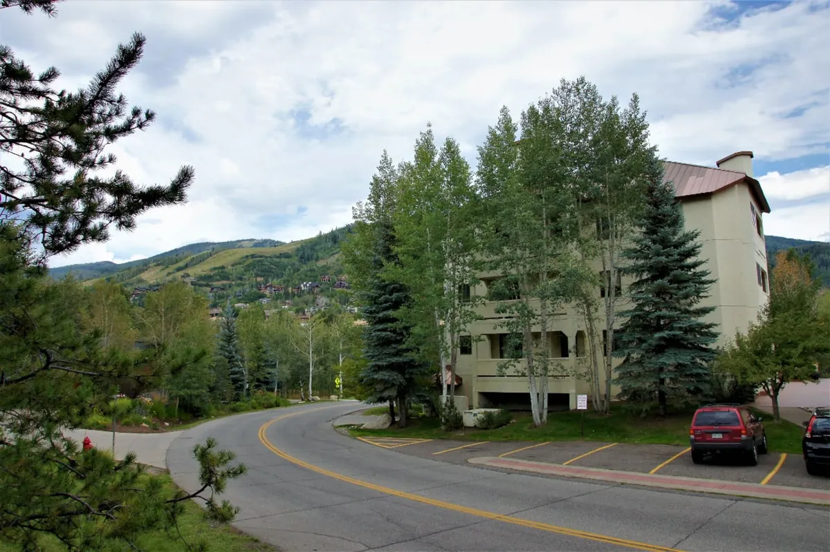 This image features the exterior of the property nestled among aspen trees, with a dramatic mountain backdrop and a curved road leading toward the building. The scene hints at immediate access to mountain adventures and the Steamboat resort area, evoking the anticipation of skiing, snowboarding, and exploration.