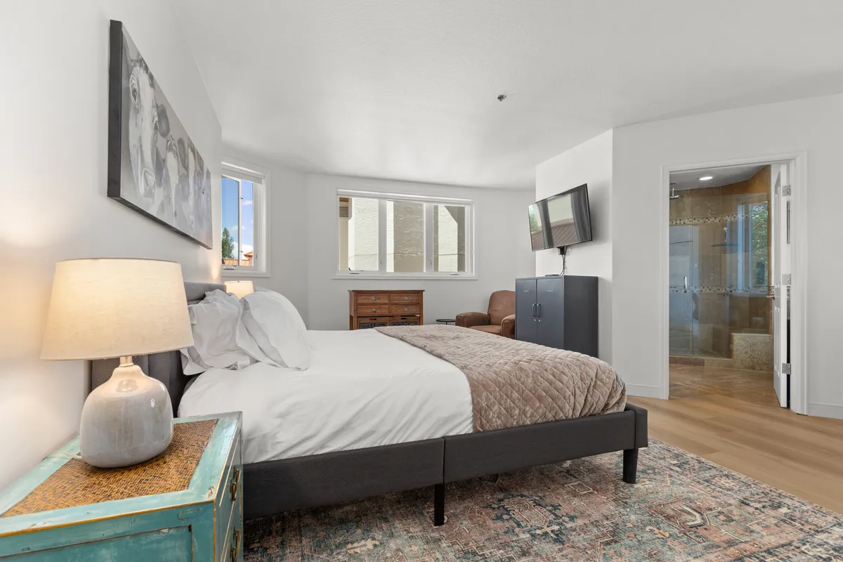 A serene bedroom featuring a plush bed with textured bedding, tasteful artwork above the headboard, soft lighting from a bedside lamp, a wooden dresser, and a wall-mounted HDTV. The space is calm and inviting, perfect for relaxation and unwinding at the end of the day.