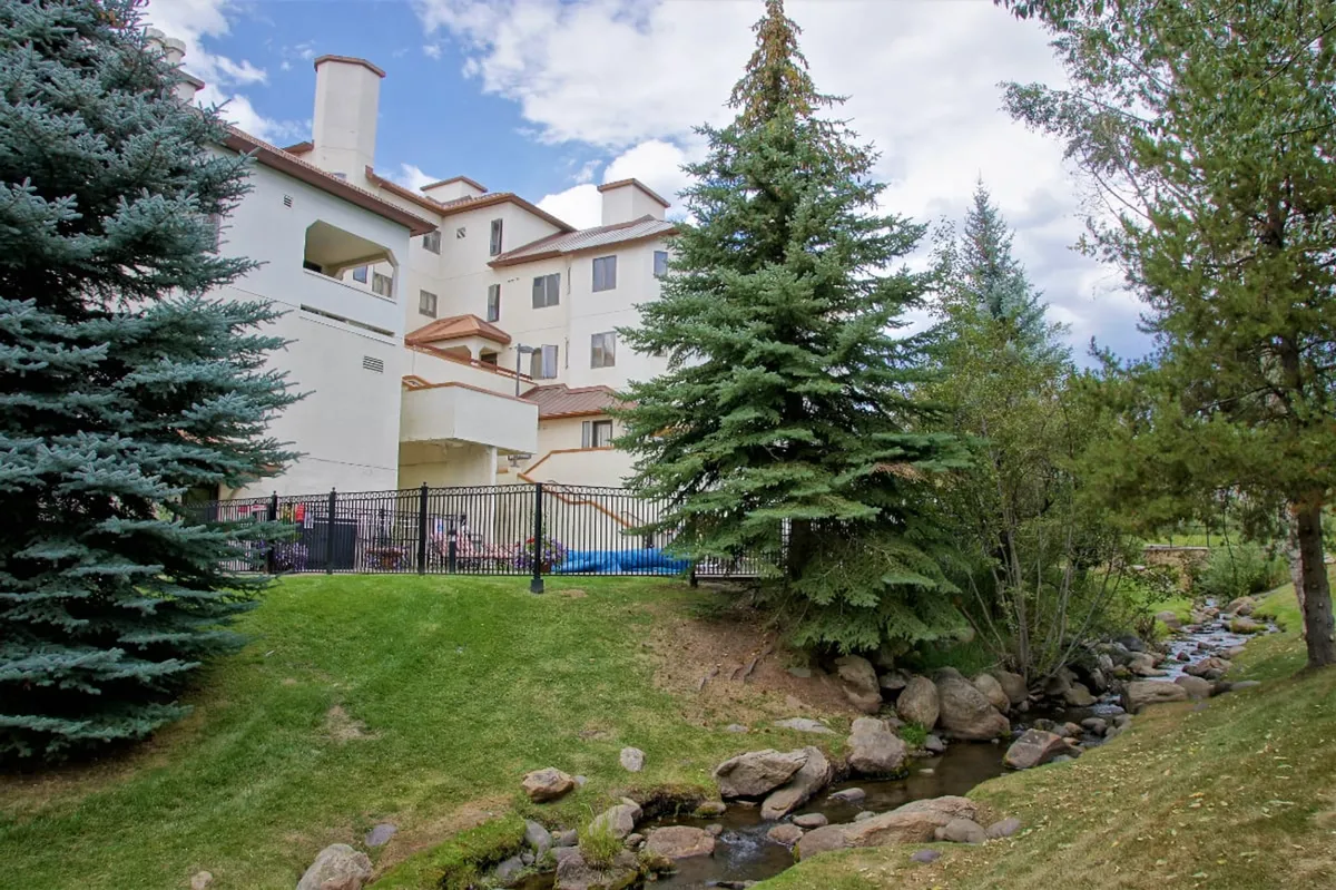 This photo shows tall evergreen trees, a gentle stream running alongside the building facade, and a welcoming patio area. The outdoor space feels integrated with the natural landscape, suggesting easy access to nature walks, streams, and opportunities for outdoor gatherings or relaxation.