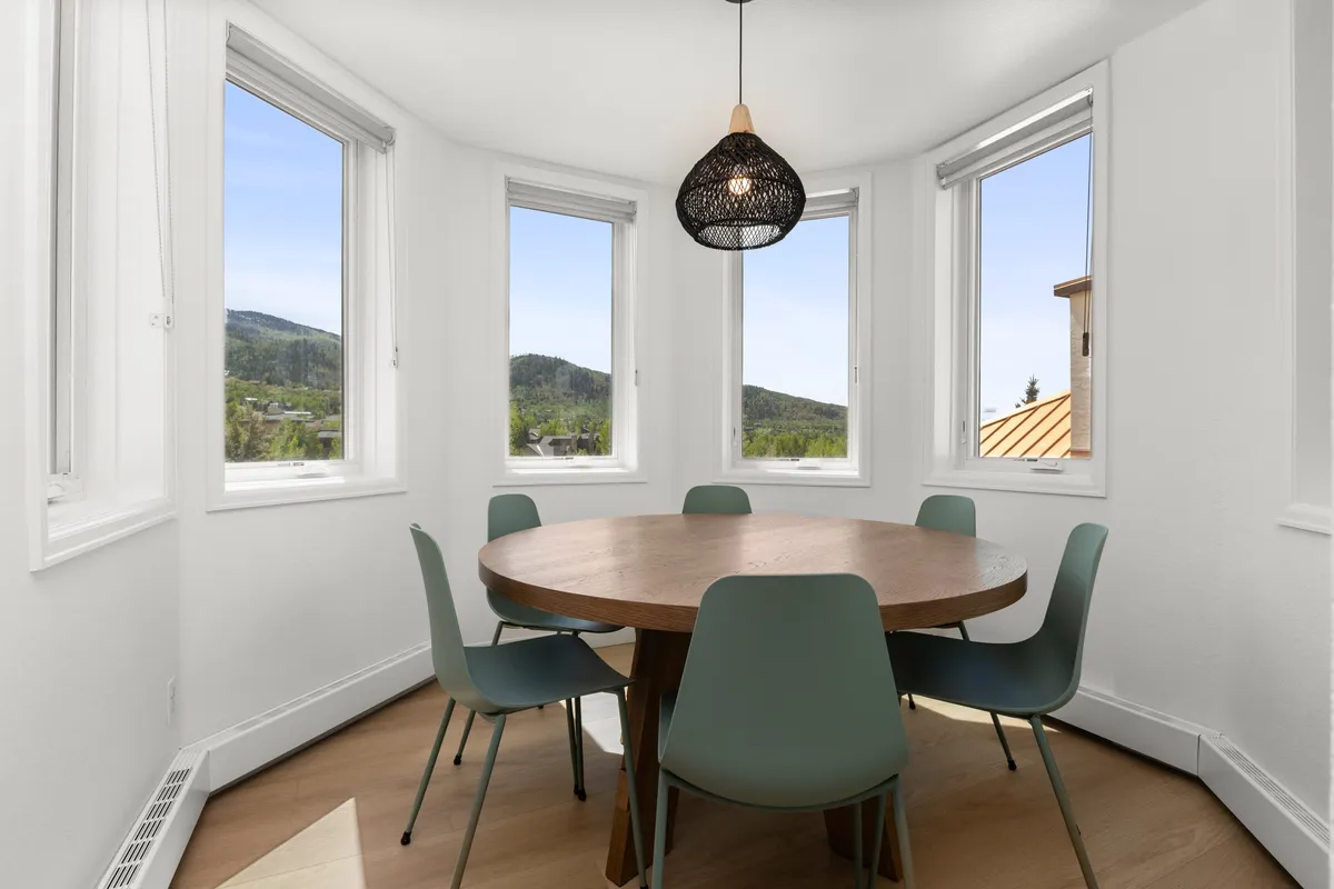 An inviting dining area with a round wooden table, green chairs, a woven pendant light, and large windows offering mountain views. The space feels cozy, perfect for intimate meals and laughter-filled gatherings with loved ones.
