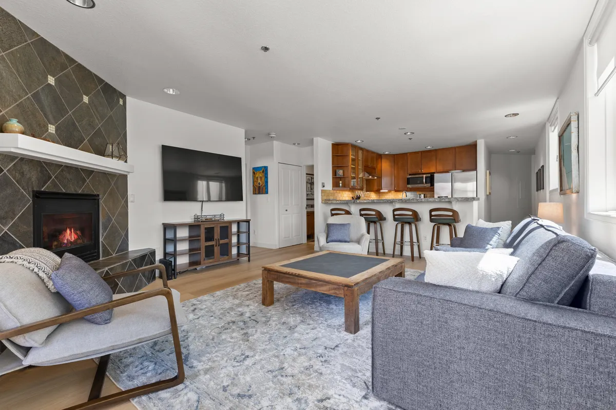 A cozy open-concept living area featuring a comfortable sofa facing a gas fireplace, with a kitchen island and bar stools visible in the background. The space is bright and inviting, with natural light streaming through the windows, and offers a seamless flow between the living, dining, and kitchen areas.