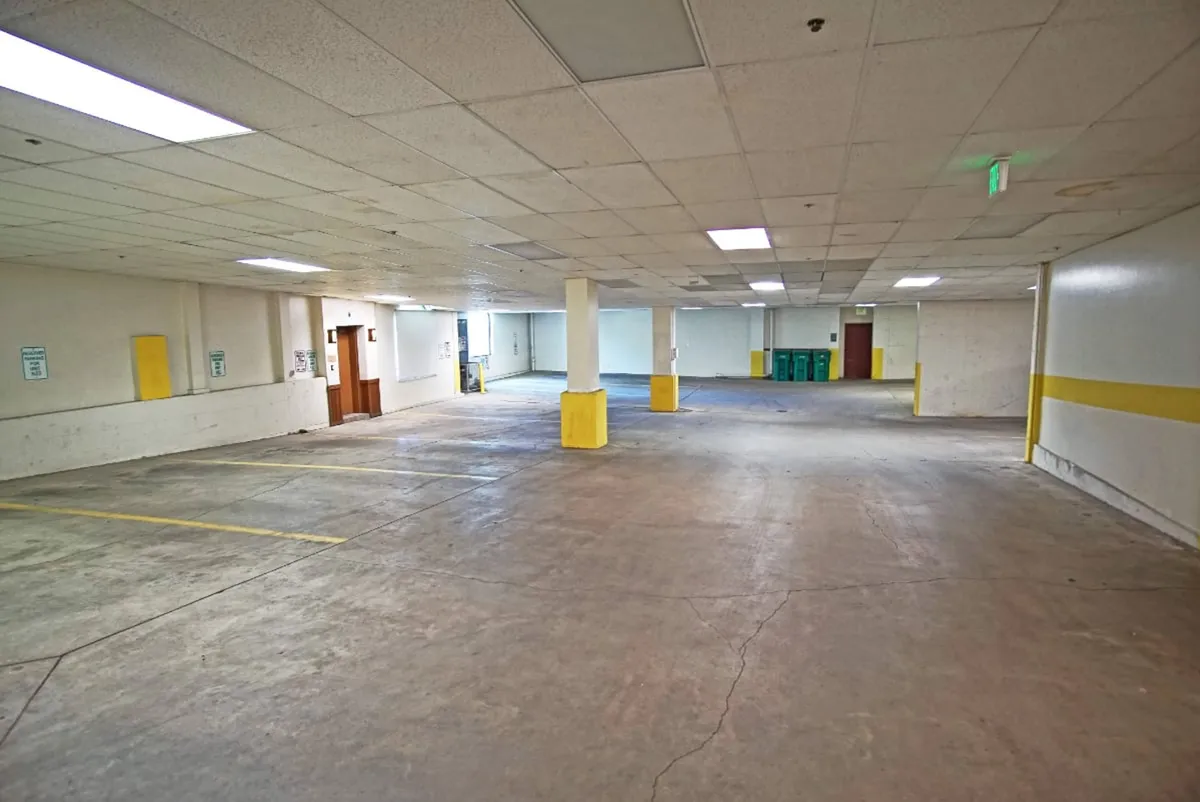 A functional indoor parking area with distinctive yellow pillars, empty parking spaces, a doorway leading into the building, and garbage bins. The scene is uncluttered and emphasizes the transition from the parking area to the interior spaces.