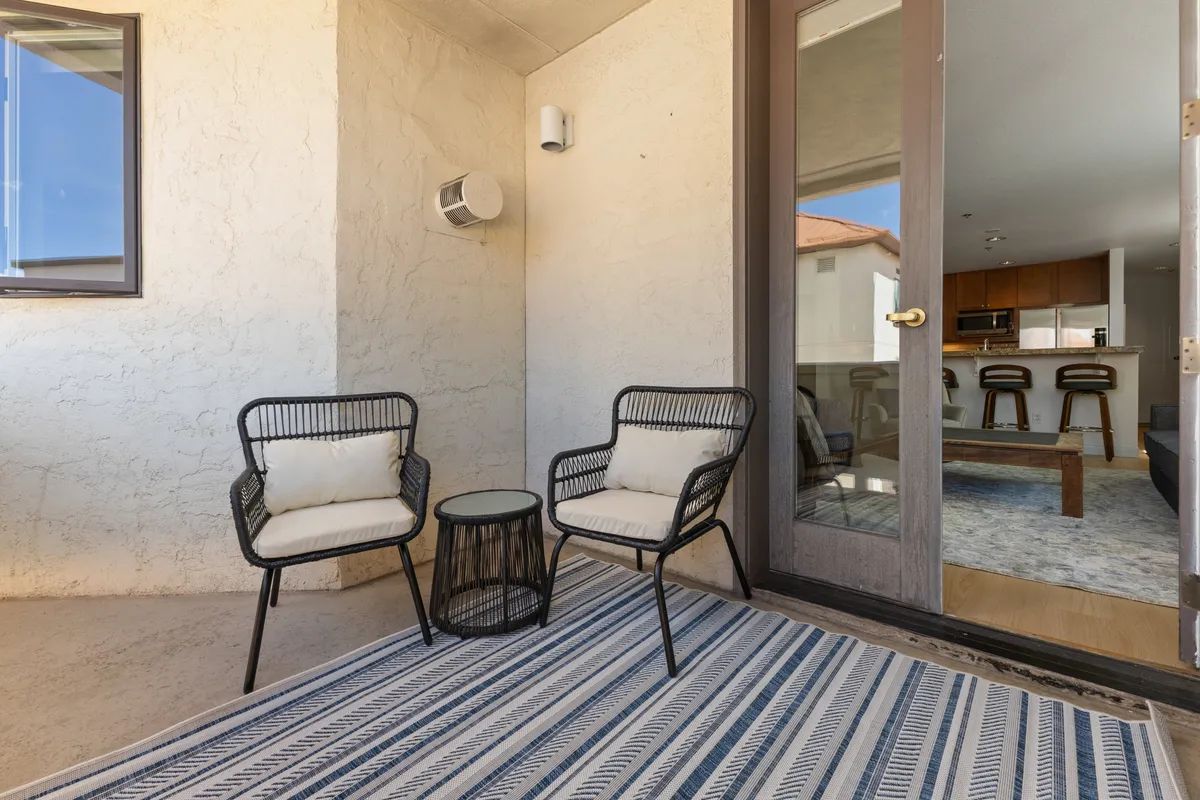 A serene covered balcony with two chairs and a small table, accented by a striped rug and a view toward nearby buildings. This space is inviting for morning coffee or an evening toast, connecting the interior living area with the outdoors.