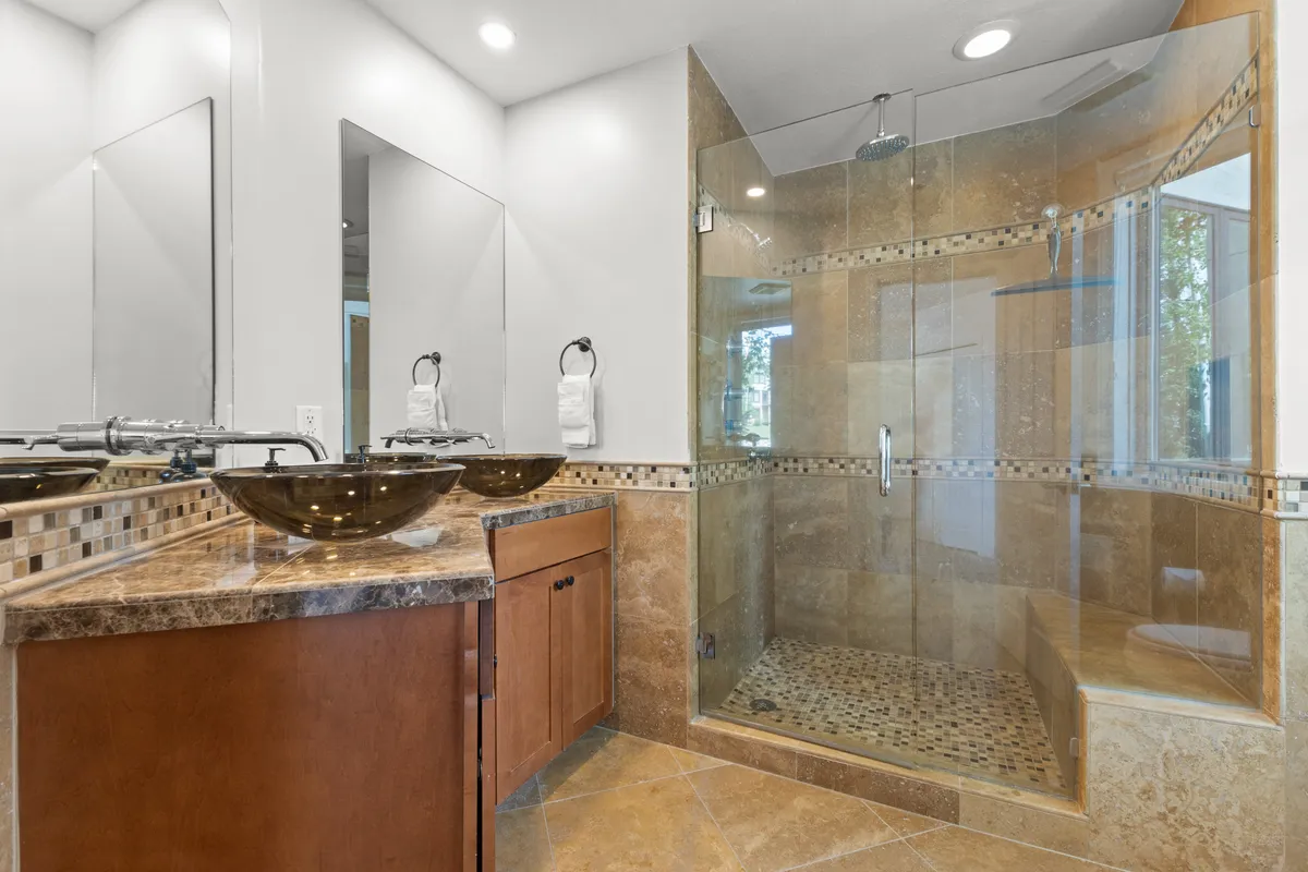 A luxurious ensuite bathroom showcasing dual vessel sinks, a spacious glass shower enclosure, and natural stone textures throughout. The space is bright and elegant, evoking a spa-like atmosphere ideal for self-care and relaxation.