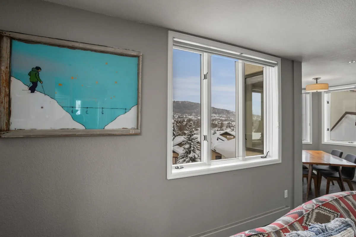 A dining nook filled with natural light from large windows, colorful artwork on the walls, and a welcoming table set for a meal. The windows offer a glimpse of the mountain landscape beyond, connecting indoor dining with scenic views.